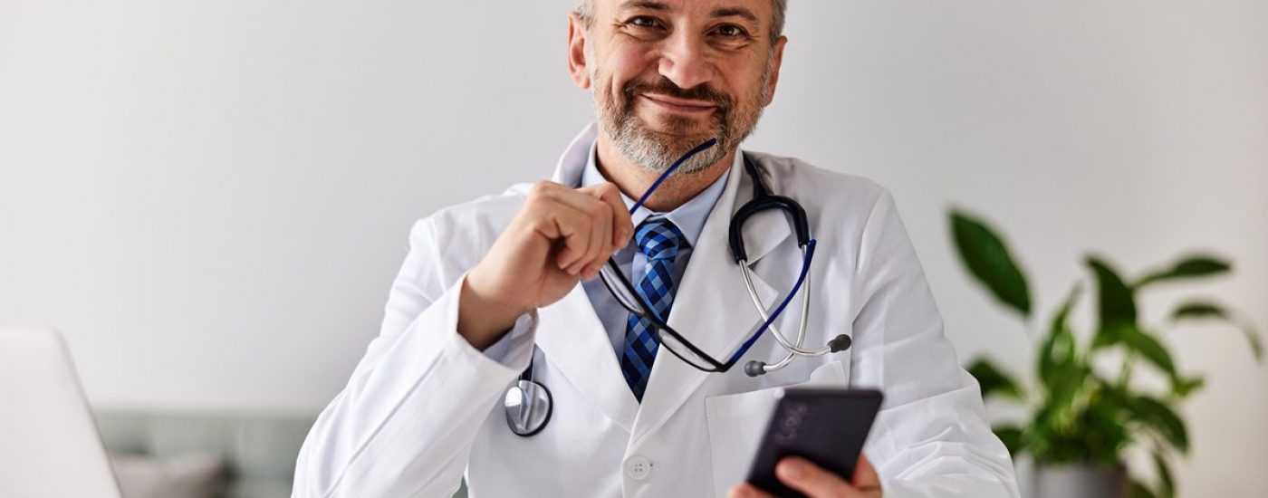 A smiling senior male doctor sitting in front of the laptop, holding a smartphone and gasses, looking at the camera.